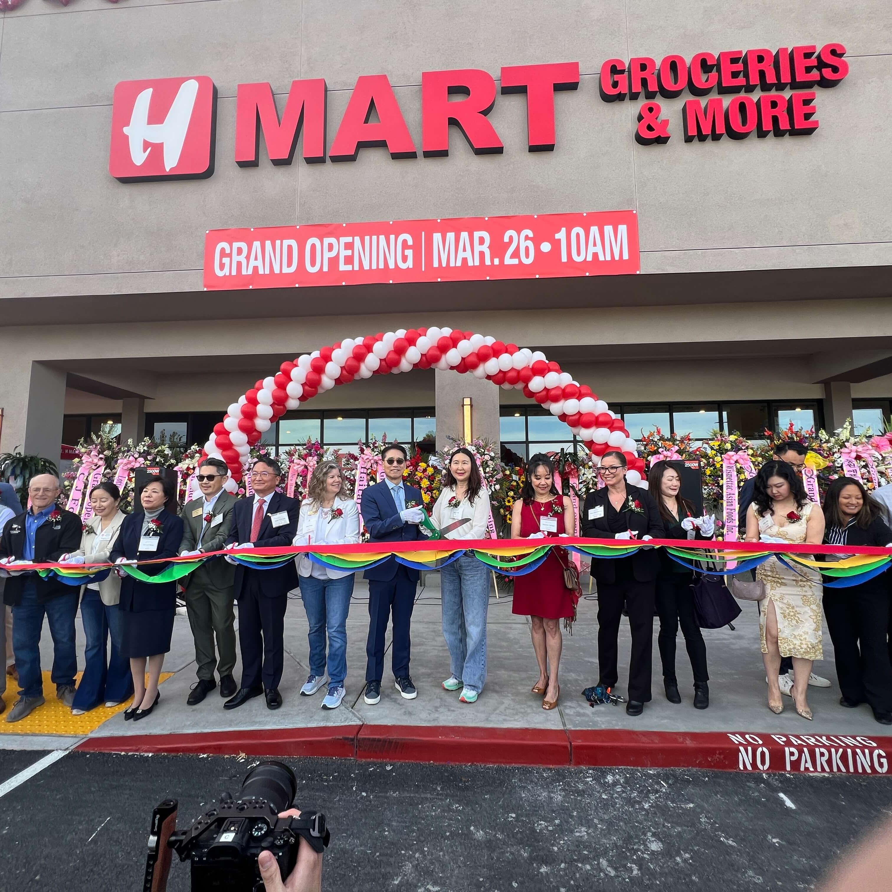 long line of people smiling and holding a rainbow colored ribbon in front of an H-Mart grocery store for a grand opening celebration